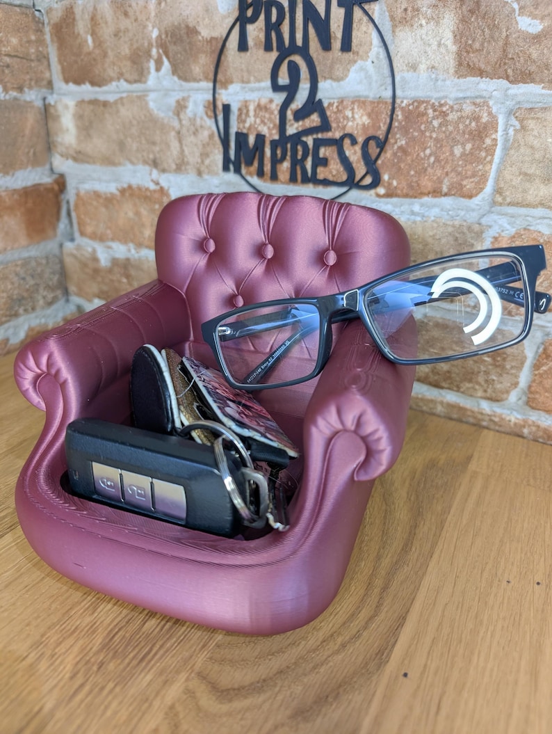 May include: A burgundy-colored miniature armchair-shaped organizer, holding a pair of black-framed eyeglasses, keys, and a small wallet. The chair has button tufting and curved armrests. The text "PRINT 2 IMPRESS" is visible in the background.