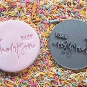 Op de afbeelding: Twee ronde, roze en grijze koekjesstempels met de tekst "BABY Thompson" en "BABY Thompson" in handschrift.