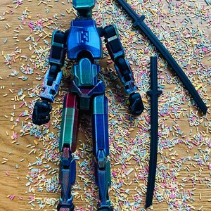 May include: A multi-colored articulated action figure with a geometric design, posed with two black katana swords. The figure is primarily blue, green, and red, with black joints. The background is covered in colorful sprinkles.