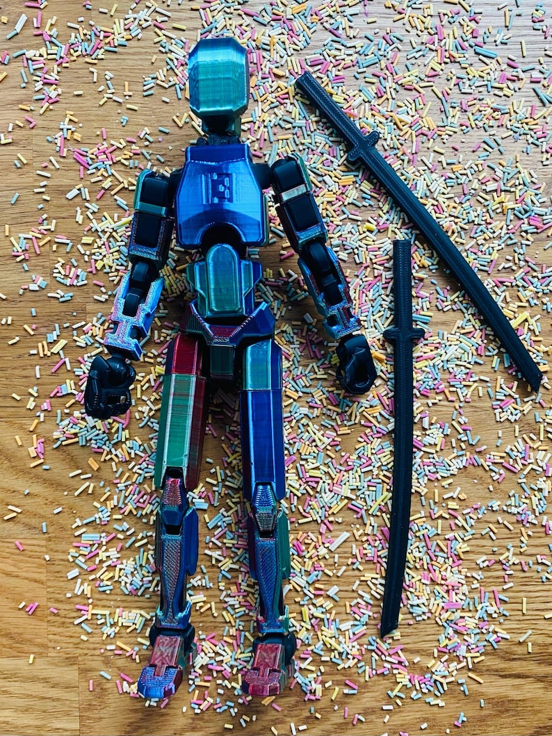 May include: A multi-colored articulated action figure with a geometric design, posed with two black katana swords. The figure is primarily blue, green, and red, with black joints. The background is covered in colorful sprinkles.