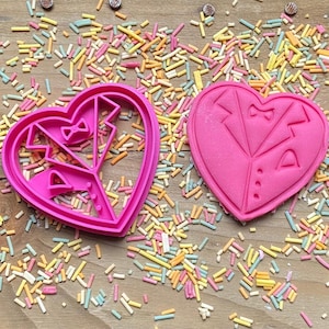 May include: Two pink heart-shaped cookie cutters, one with a detailed outline of a tuxedo and the other with a solid tuxedo design.