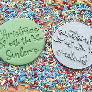 May include: Two round green and gray cookie cutters with the text "Christmas with the Sinlers" and "Santa with the Sinlers" on them. The cookie cutters are surrounded by colorful sprinkles.