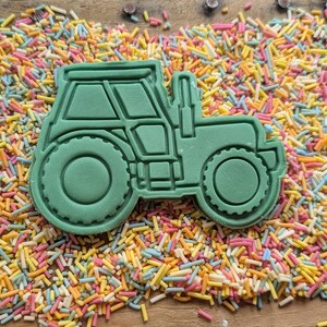 May include: A green tractor-shaped cookie cutter with detailed windows, wheels, and body. The cutter is surrounded by colourful sprinkles on a wooden surface. Ideal for baking themed treats.