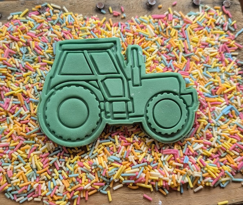 May include: A green tractor-shaped cookie cutter with detailed windows, wheels, and body. The cutter is surrounded by colourful sprinkles on a wooden surface. Ideal for baking themed treats.
