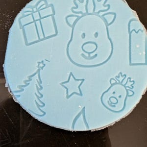 May include: A light blue, circular mold with Christmas-themed designs. The mold features embossed images of a gift box, a Christmas tree, a star, a reindeer head, and a mountain. The mold is likely used for crafting or baking.