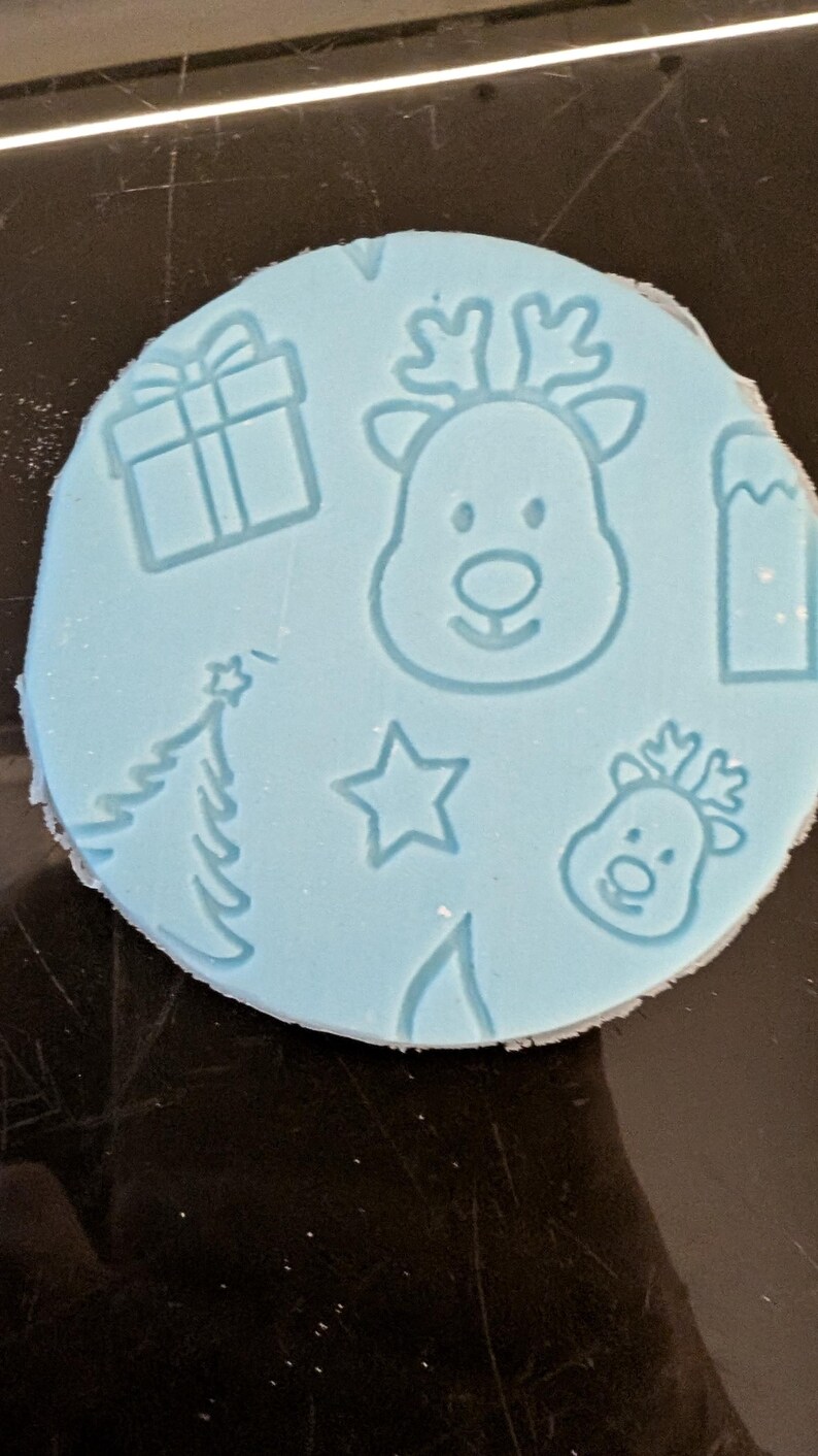 May include: A light blue, circular mold with Christmas-themed designs. The mold features embossed images of a gift box, a Christmas tree, a star, a reindeer head, and a mountain. The mold is likely used for crafting or baking.