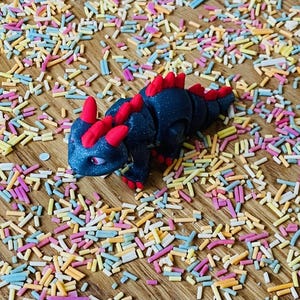 May include: A black and red articulated toy dragon with a glittery finish. The dragon is posed on a wooden surface covered in colorful sprinkles.