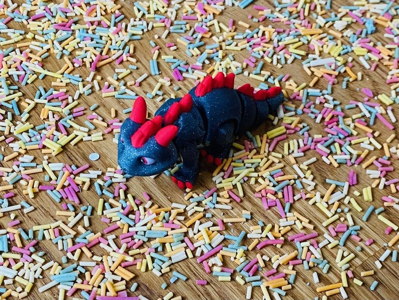 May include: A black and red articulated toy dragon with a glittery finish. The dragon is posed on a wooden surface covered in colorful sprinkles.
