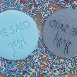 May include: Two round, blue and gray, silicone stamps with the text "She Said Yes!" in white lettering. The stamps are on a background of colorful sprinkles.