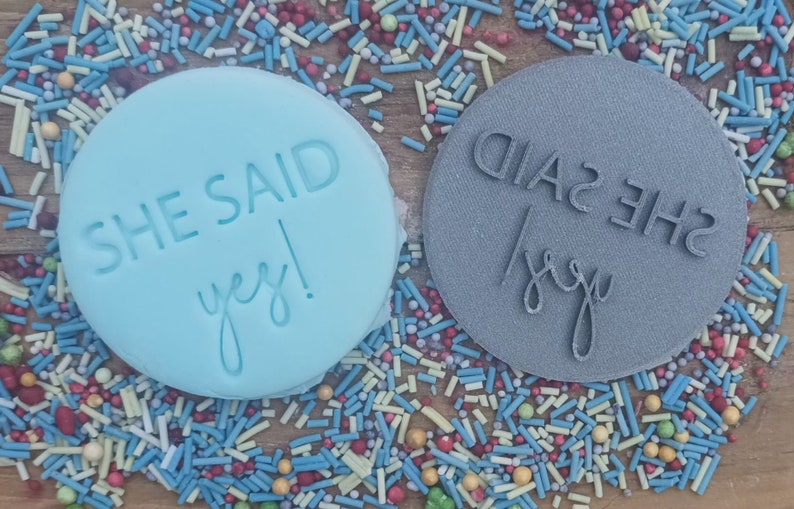 May include: Two round, blue and gray, silicone stamps with the text "She Said Yes!" in white lettering. The stamps are on a background of colorful sprinkles.
