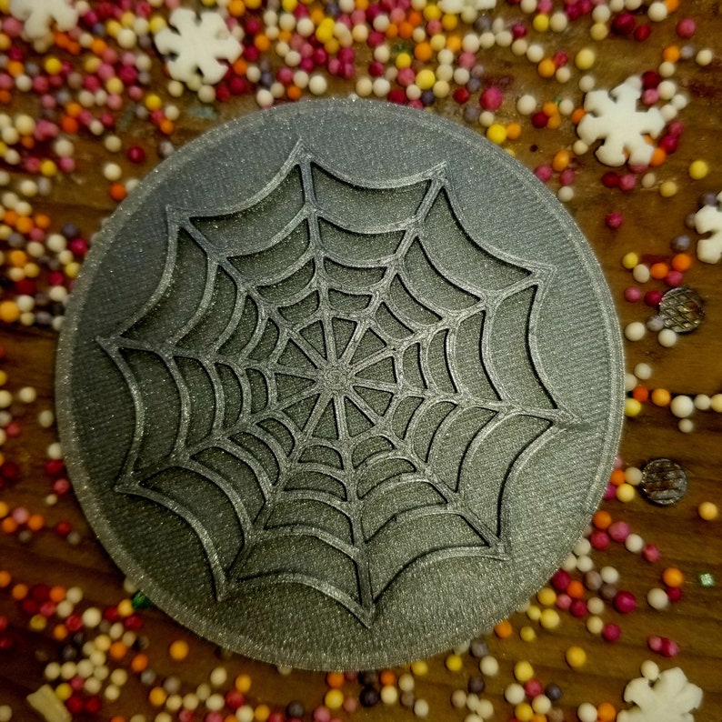 May include: A gray, round, 3D printed cookie cutter with a spiderweb design. The cookie cutter is made of a textured material and has a shiny finish.