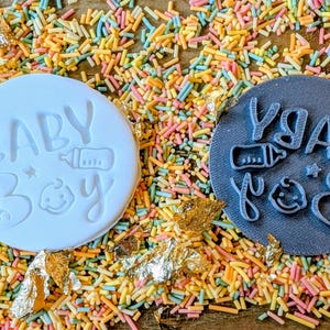 May include: A white circular stamp with the words "BABY Boy" and a baby bottle and a smiling baby face. Next to it is a dark gray stamp with the same design. The stamps are on a bed of colorful sprinkles.
