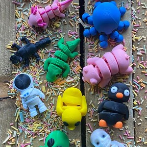 May include: A collection of 15 colorful, 3D printed figurines, including a turtle, astronaut, octopus, pig, penguin, alien, and skeleton. The figurines are arranged on a surface covered in colorful sprinkles.