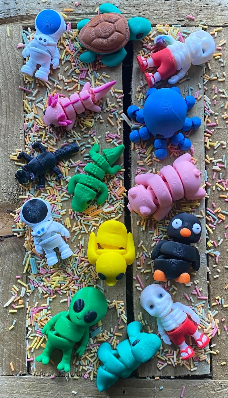 May include: A collection of 15 colorful, 3D printed figurines, including a turtle, astronaut, octopus, pig, penguin, alien, and skeleton. The figurines are arranged on a surface covered in colorful sprinkles.