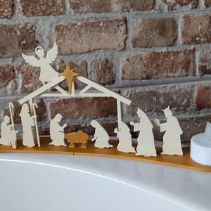 May include: A wooden nativity scene with an angel, star, and figures of the Holy Family and the Three Wise Men. The scene is set on a curved wooden base, with two battery-operated tea lights on either end. The background is a brick wall.