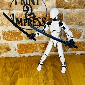 May include: A white articulated action figure with black joints and two black swords. The figure is posed in a fighting stance. The background includes a brick wall and a sign that reads "PRINT 2 IMPRESS".