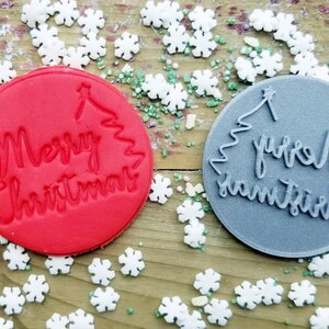 May include: Two circular cookie stamps with the words "Merry Christmas" and a Christmas tree design. One stamp is red, the other is gray. White snowflake sprinkles and green sprinkles surround the stamps on a wooden surface.