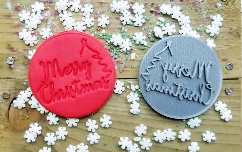 May include: Two circular cookie stamps with the words "Merry Christmas" and a Christmas tree design. One stamp is red, the other is gray. White snowflake sprinkles and green sprinkles surround the stamps on a wooden surface.
