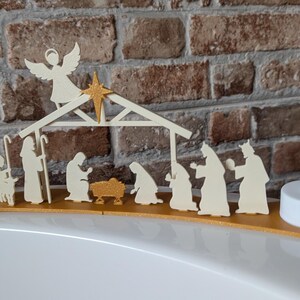 May include: A wooden nativity scene with an angel, star, and figures of Mary, Joseph, baby Jesus, and the Three Wise Men. The scene is set on a gold-colored base, flanked by two white LED candles. The background is a brick wall.