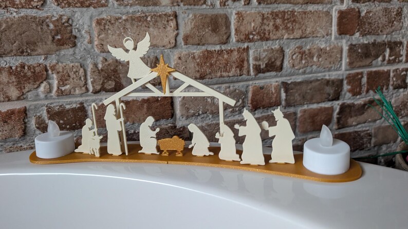 May include: A wooden nativity scene with an angel, star, and figures of Mary, Joseph, baby Jesus, and the Three Wise Men. The scene is set on a gold-colored base, flanked by two white LED candles. The background is a brick wall.