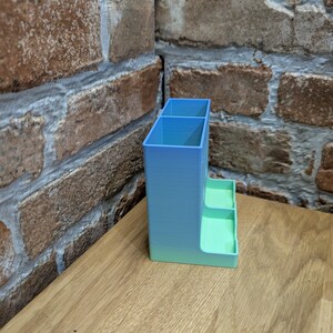 May include: A 3D-printed desk organizer in a gradient of blue and green. The organizer has two compartments and two small trays. It is placed on a wooden surface, with a brick wall in the background.