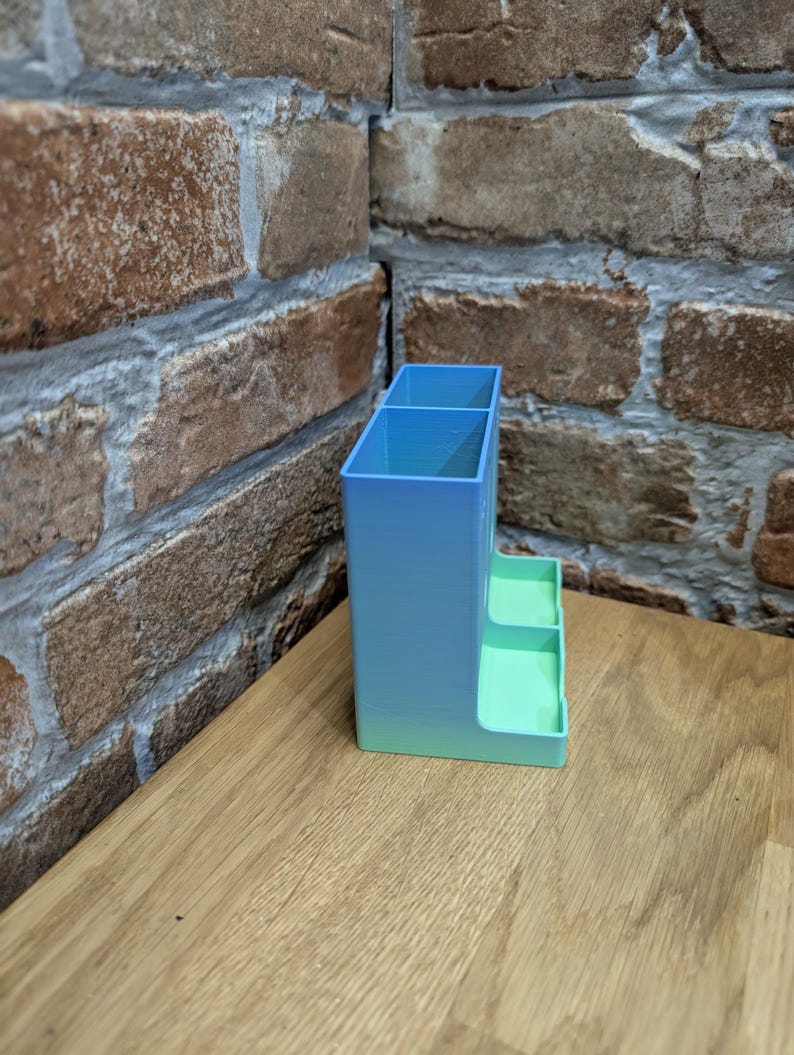 May include: A 3D-printed desk organizer in a gradient of blue and green. The organizer has two compartments and two small trays. It is placed on a wooden surface, with a brick wall in the background.