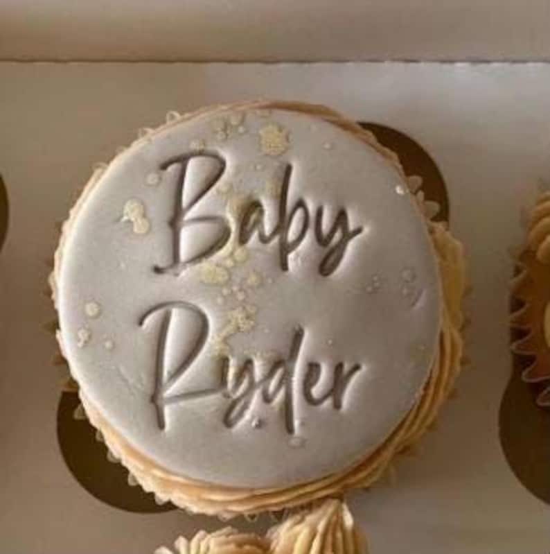 May include: A frosted cupcake with the words "Baby Ryder" in elegant script. The frosting is light gray with gold flecks, and the cupcake sits in a decorative paper cup. The overall theme suggests a baby shower or celebration.