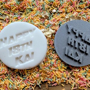 May include: A white, round edible cake decoration with the words "HAPPY 18TH KAI" embossed on it, next to a gray, round stamp with the same text in reverse. The items are surrounded by colorful sprinkles.