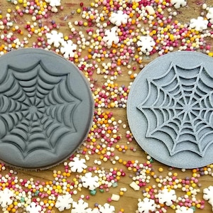 May include: Two gray plastic cookie cutters shaped like spiderwebs. The cutters are on a wooden surface with colorful sprinkles.