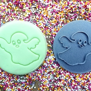 May include: Two cookie cutters, one green and one gray, shaped like a ghost with a cute face. The cutters are on a background of colorful sprinkles.