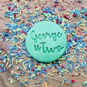 May include: A green round cookie cutter with the text "George is Two" in white lettering. The cookie cutter is surrounded by colorful sprinkles.