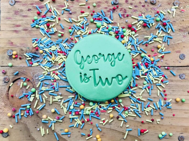 May include: A green round cookie cutter with the text "George is Two" in white lettering. The cookie cutter is surrounded by colorful sprinkles.