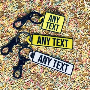 May include: Three black keychains with yellow and white rectangular tags that say "ANY TEXT". The keychains are on a surface covered in colorful sprinkles.