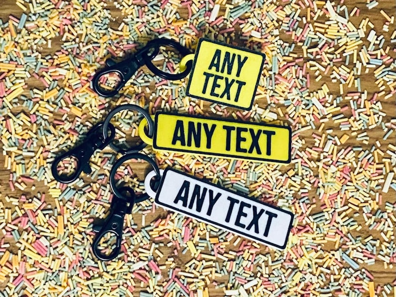 May include: Three black keychains with yellow and white rectangular tags that say "ANY TEXT". The keychains are on a surface covered in colorful sprinkles.