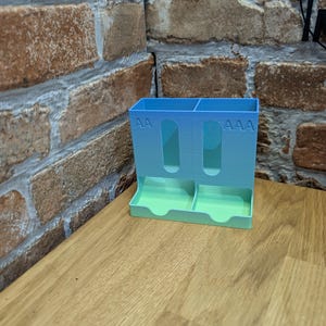 May include: A 3D-printed battery organizer in a gradient of blue and green. The organizer has slots for AA and AAA batteries, with the corresponding text printed on the front. It sits on a wooden surface.