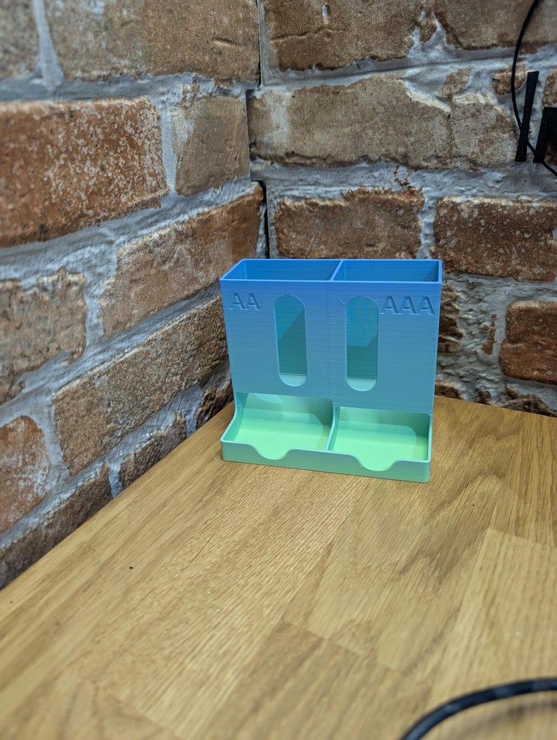 May include: A 3D-printed battery organizer in a gradient of blue and green. The organizer has slots for AA and AAA batteries, with the corresponding text printed on the front. It sits on a wooden surface.