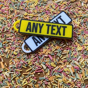 May include: Two yellow and white rectangular keychains with the text "ANY TEXT" printed in black. The keychains are on a background of colorful sprinkles.