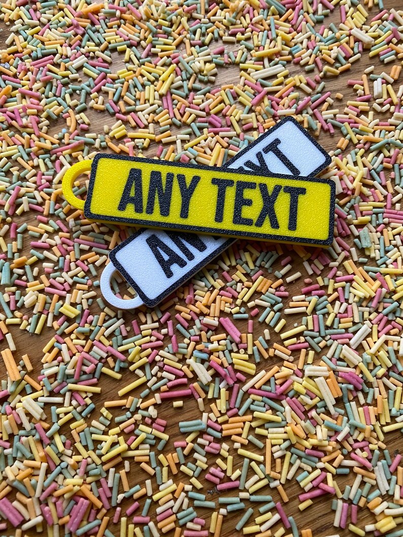 May include: Two yellow and white rectangular keychains with the text "ANY TEXT" printed in black. The keychains are on a background of colorful sprinkles.