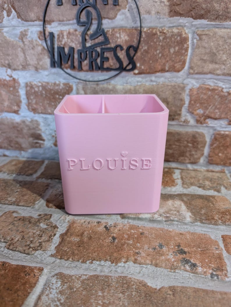May include: A light pink, square-shaped desk organizer with two compartments. The word "PLOUISE" is embossed on the front. The background features a brick wall and a black circular sign that says "IMPRESS".