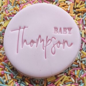 Op de afbeelding: Een ronde roze koek met de tekst "BABY Thompson" in roze letters.