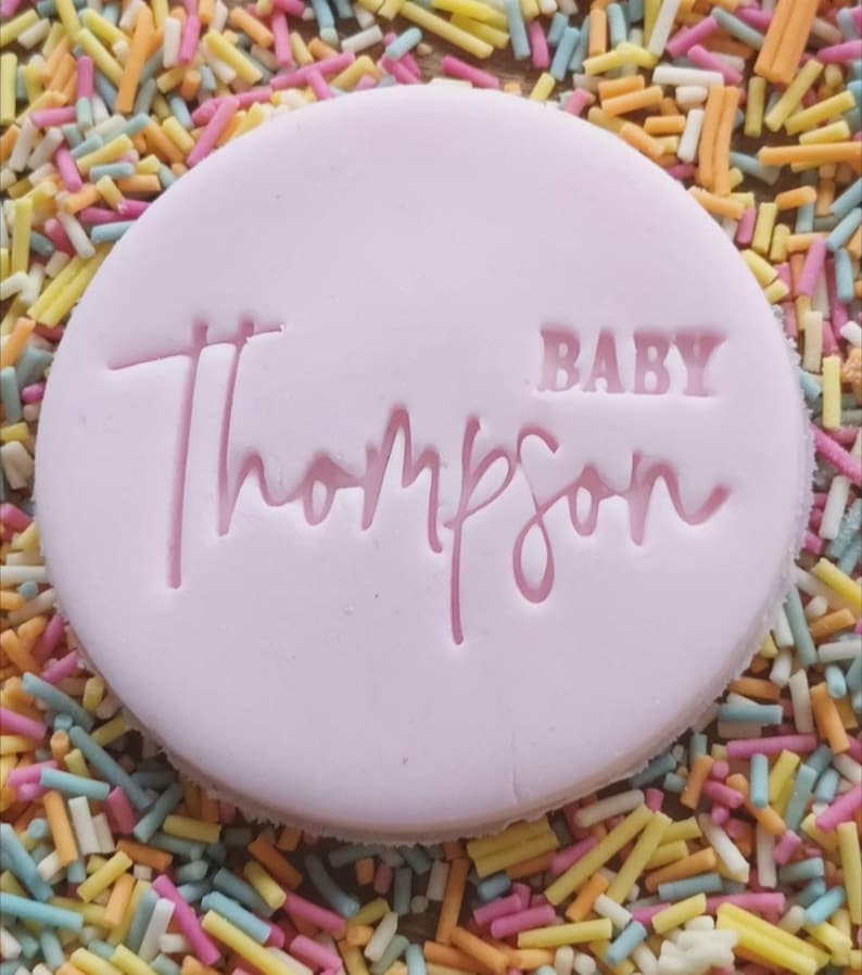 Op de afbeelding: Een ronde roze koek met de tekst "BABY Thompson" in roze letters.