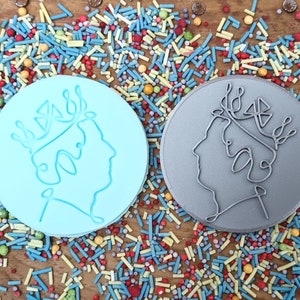 May include: Two circular cookie cutters, one light blue and one gray, each featuring a profile of a crowned head. The cutters are surrounded by colorful sprinkles on a wooden surface. The design is a line drawing.