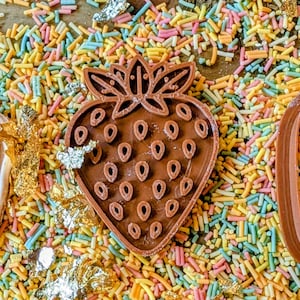 May include: Three strawberry-shaped items on a bed of colorful sprinkles. A light pink strawberry-shaped cookie cutter, a brown chocolate strawberry, and a brown strawberry-shaped cookie cutter. Gold leaf flakes are scattered around the items.