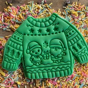 K&ouml;nnte beinhalten: Ein gr&uuml;ner Keks in Form eines Weihnachtspullovers, verziert mit Sternen, B&auml;umen und zwei Figuren mit Weihnachtsm&uuml;tzen. Der Keks liegt auf einer Holzoberfl&auml;che, die mit bunten Streuseln bedeckt ist.