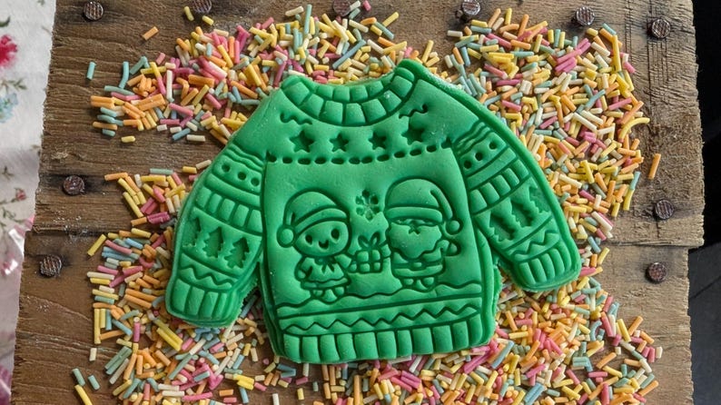 K&ouml;nnte beinhalten: Ein gr&uuml;ner Keks in Form eines Weihnachtspullovers, verziert mit Sternen, B&auml;umen und zwei Figuren mit Weihnachtsm&uuml;tzen. Der Keks liegt auf einer Holzoberfl&auml;che, die mit bunten Streuseln bedeckt ist.