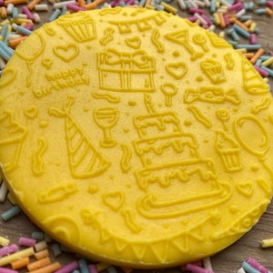 May include: A yellow, circular item with embossed birthday-themed designs. The design includes a three-tiered cake, party hat, gift, and the words "happy birthday". Colorful sprinkles surround the item.