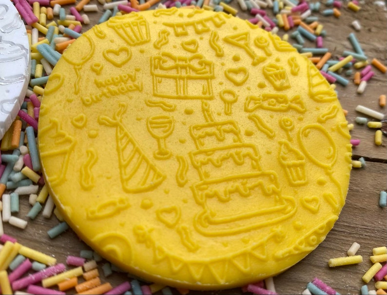 May include: A yellow, circular item with embossed birthday-themed designs. The design includes a three-tiered cake, party hat, gift, and the words "happy birthday". Colorful sprinkles surround the item.
