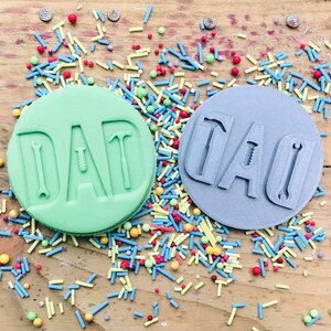 Pu&ograve; includere: Due timbri rotondi per biscotti, verde e grigio, con la scritta "DAD" e disegni di attrezzi. I timbri sono circondati da zuccherini colorati su una superficie di legno.