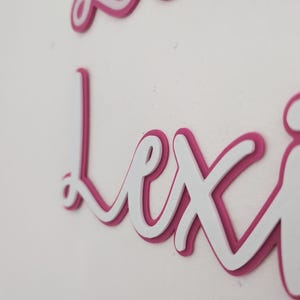 May include: A white and pink layered name sign with the name "Lexi" in cursive script.