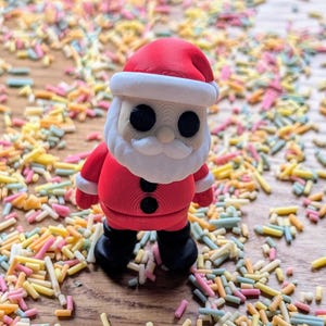 Op de afbeelding: Een rode en witte kerstmanfiguur met een zwarte knoop op zijn jas, staand op een oppervlak bedekt met kleurrijke sprinkels.